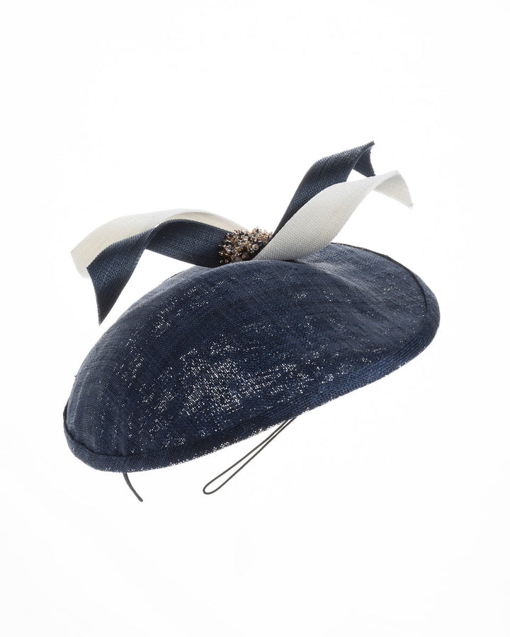 Still ife picture of an asymmetric navy saucer, decorated with white and navy buntal swirls and completed with multi coloured crystal beads. The height gained on the left side of this headpiece creates a dramatic effect and completes your style with a unique touch. Designed and made by Merve Bayindir millinery.