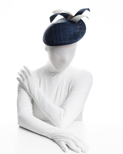 Model is showcasing an asymmetric navy saucer, decorated with white and navy buntal swirls and completed with multi colored crystal beads. The height gained on the left side of this headpiece creates a dramatic effect and completes your style with a unique touch. The hat has a higher side on the left side.