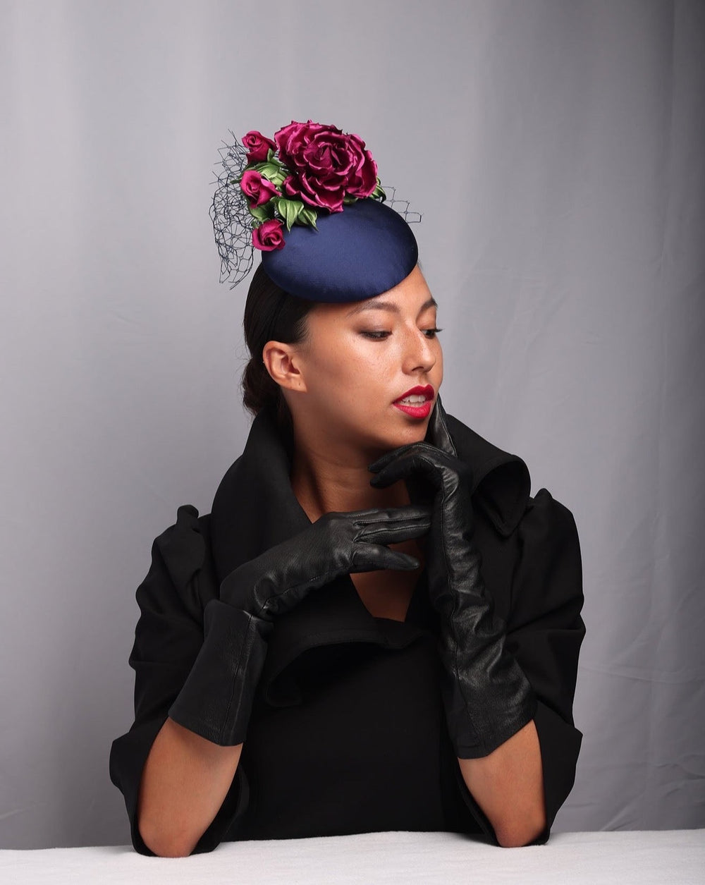 The mode is showcasing the side of a Sateen button shaped fascinator, decorated with dark pink sateen roses embellished with green leaves and navy veil.