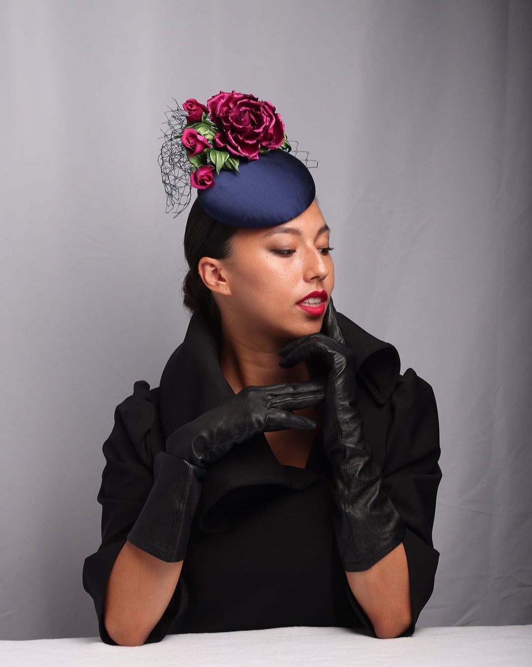 The mode is showcasing the side of a Sateen button shaped fascinator, decorated with dark pink sateen roses embellished with green leaves and navy veil.