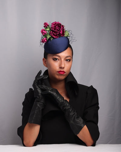 The model is showcasing the front view of a Sateen button shaped fascinator, decorated with dark pink sateen roses embellished with green leaves and navy veil.