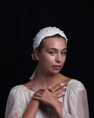 elegant white satin headband features delicate pearl-like embellishments scattered across its surface, adding a touch of glamour. A wide satin ribbon forms a neat bow at the back, with the ribbon elegantly hanging down. The padded structure ensures a comfortable and secure fit. Perfect for weddings or formal events, this headband exudes refined sophistication.