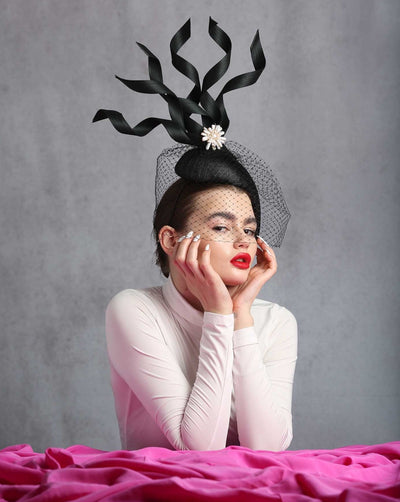 Model is showcasing the side of a Tear drop shaped black sinamay base fascinator decorated with jinsin swirls rising towards up, all attached to gether with mother of pearl flower. Fascinator is embellished with a full veil surrounding the fascinator.