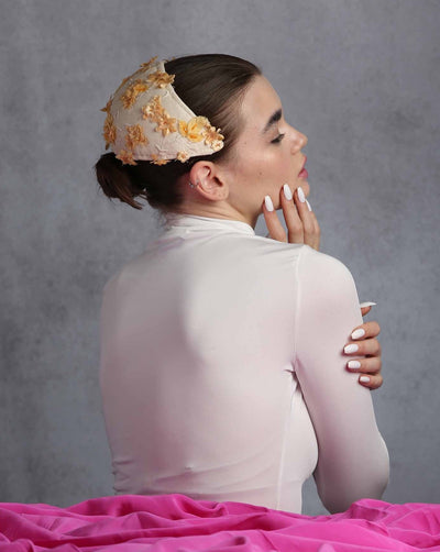 Model is showcasing the back of a vintage-style half hat features a cream fabric adorned with delicate beige and yellow embroidered flowers and tiny bead accents. It is designed to sit comfortably on the head, curving gracefully to frame the back and sides. The hat’s intricate detailing and subtle color palette add a touch of classic elegance