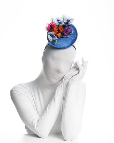 Model is showcasing the front view of a deep blue saucer style mini fascinator, decorate with different coloured violet flowers.