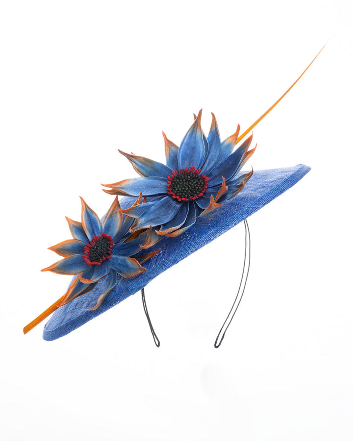Deep blue disk style wide brim hat, decorated with blue sunflower style flowers with orange edges, its embellished with an orange coloured ostrich quill. Designed and made by Merve Bayindir Millinery.

