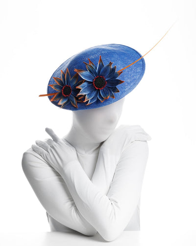 The model is showcasing the top view of a Deep blue disk style wide brim hat, decorated with blue sunflower style flowers with orange edges, its embellished with an orange coloured ostrich quill.