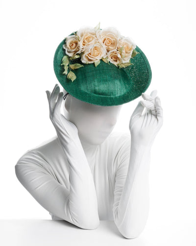 Model is showcasing a Medium sized emerald green saucer hat decorated with a bouquet on top back side. The bouquet features six flowers in different sizes and shades ranging from light peach to yellow.