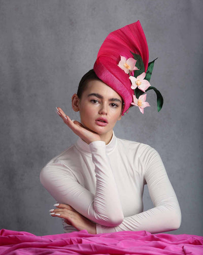 The model is showcasing a Pink flat tear drop shaped fascinator. highten by a silk abaca fan like back curved at the back of the fascinator. The bottom edge of the fascinator decorated with light pink orchids and green leaves.