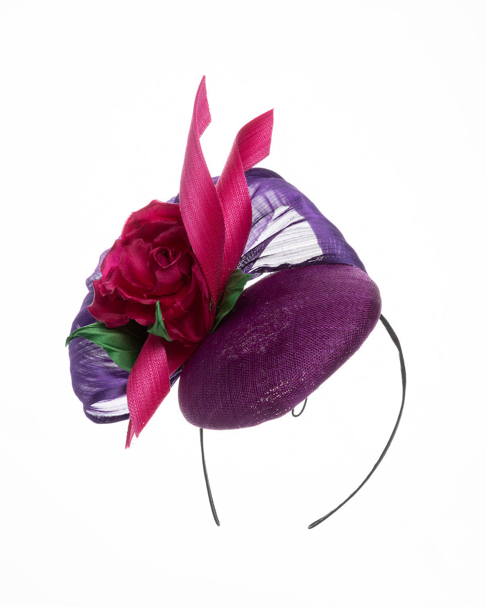 A purple sinamay button shaped fascinator decorated with curved silk abaca fabric curving like a sea shell, two buntal swirls and a large pink silk rose, exemplifying the harmony of different shades of color. Designed and made by Merve Bayindir Millinery.
