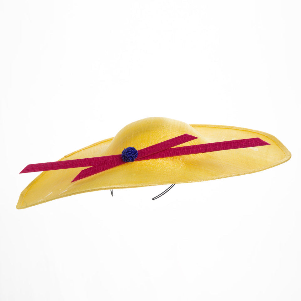 Still life picture of wide brim asymmetric yellow hat tilted to the left. Decorated with two pink buntal stripes crossing the hat as well as each other, embellished the centre of straight ribbon is a dark blue bead bead cluster. Designed and made by Merve Bayindir millinery.
