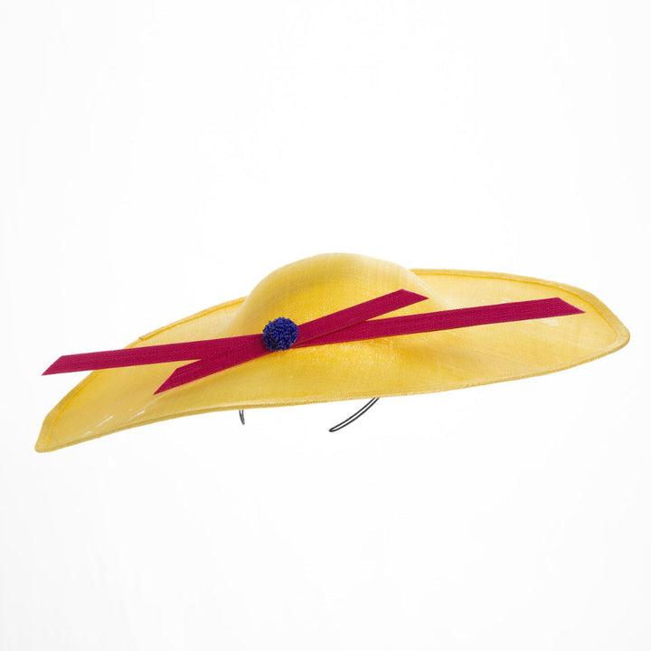 Still life picture of wide brim asymmetric yellow hat tilted to the left. Decorated with two pink buntal stripes crossing the hat as well as each other, embellished the centre of straight ribbon is a dark blue bead bead cluster. Designed and made by Merve Bayindir millinery.