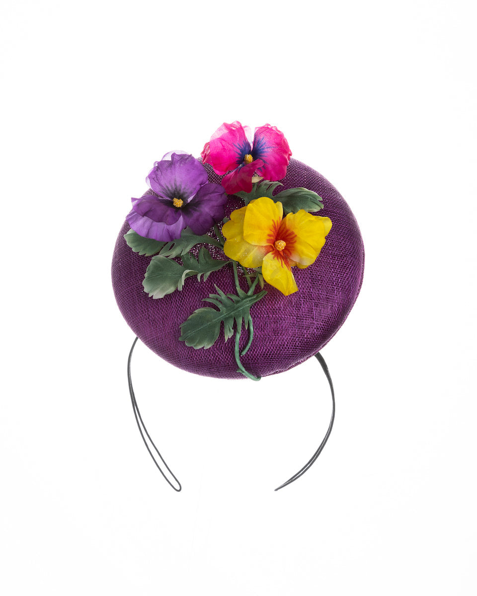 Purple button shape fascinator, that sirs on the centre of the head, the fascinator is decorated with a purplue and a pink and a yellow violet bouquet with stems and leaves. Designed and made by Merve Bayindir Millinery