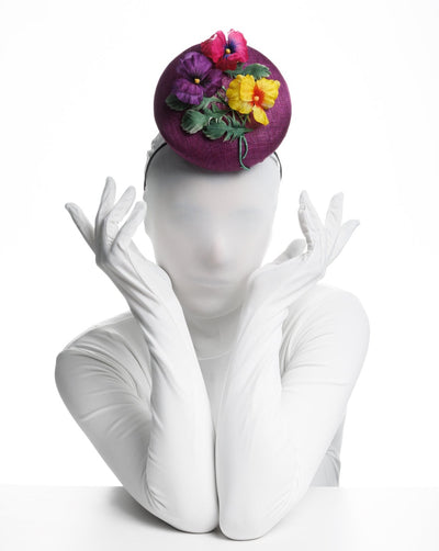 Model is showcasing the front view of a  Purple button shape fascinator, that sirs on the centre of the head, the fascinator is decorated with a purplue and a pink and a yellow violet bouquet with stems and leaves.