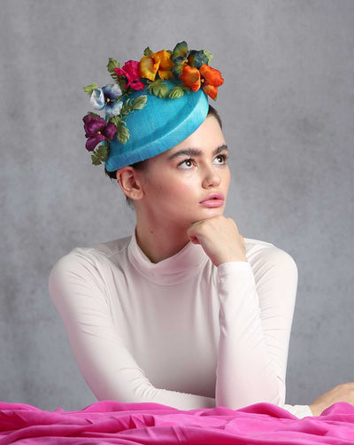 Model is showcasing the right side of a Asymmetric preacher style teal coloured Sinamay fascinator decorated with custom made silk organza multi-colored violets.