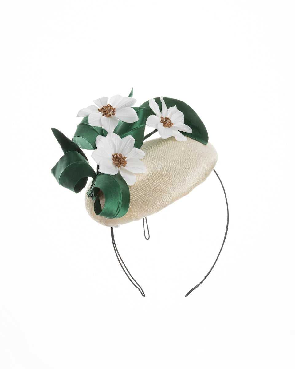 Val is a button shaped, ivory sinamay fascinator worn to the front right side of the head. The fascinator is decorated with delicate white silk flowers and green sateen leaf swirls. Designed and made by Merve Bayindir millinery.