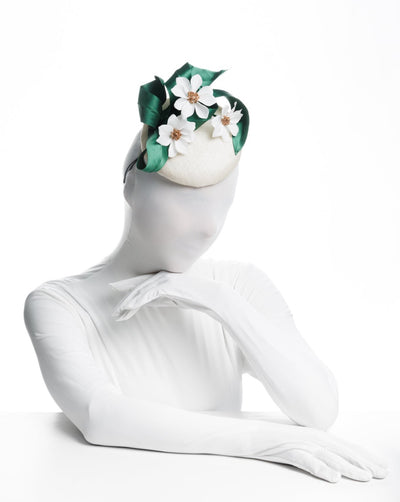 Model is showcasing the front view of a button shaped, ivory sinamay fascinator worn to the front right side of the head. The fascinator is decorated with delicate white silk flowers and green sateen leaf swirls.