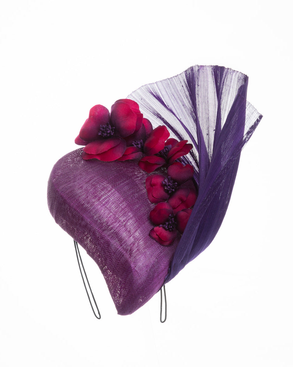 West is a purple side percher style fascinator curving to the left of the head framing the right side of the face. Decorated with a variety of pink and purple shaded cherry flowers and completed with a purple pleaded fan in the back. Designed and made by Merve Bayindir Millinery.
