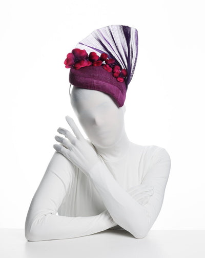 Model is showcasing the front view of a purple side percher style fascinator curving to the left of the head framing the right side of the face. Decorated with a variety of pink and purple shaded cherry flowers and completed with a purple pleaded fan in the back.