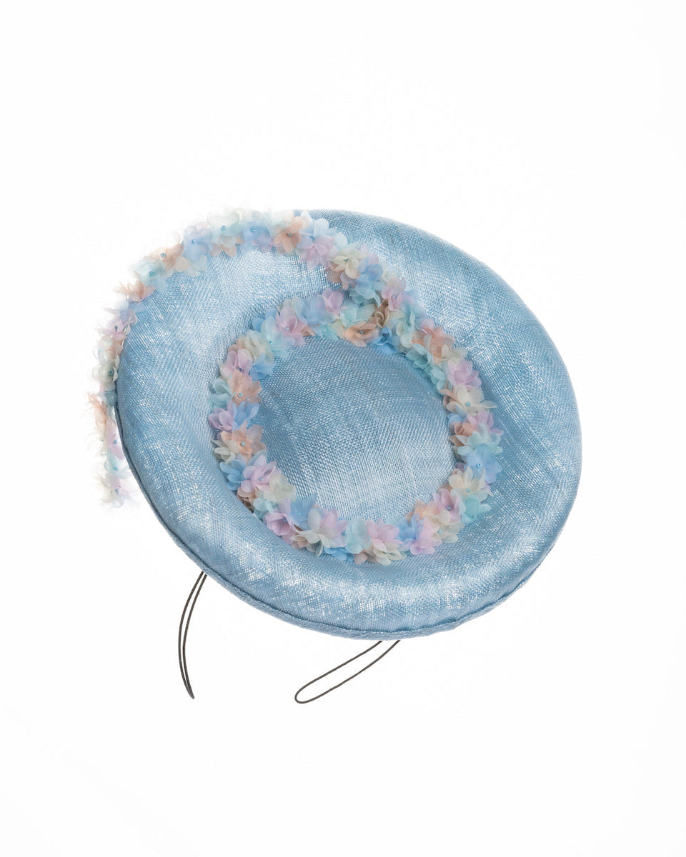  A light blue, curved oval shaped high fascinator decorated with pastel colored organza flowers that circle the center and extend towards the back, providing additional flare. Designed and made by Merve Bayindir Millinery.
