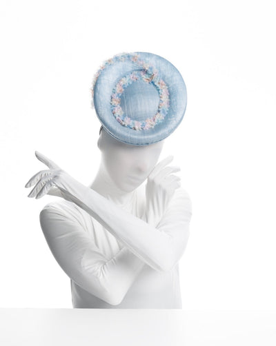 Model is showcasing  a light blue, curved oval shaped high fascinator decorated with pastel colored organza flowers that circle the center and extend towards the back, providing additional flare. The fascinator sits on the right front side of the head.