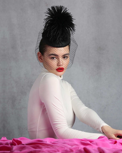 The model is showcasing the front view of a black fascinator. lack button shaped fascinator decorated with feather pompon. The full surrounding veil is decorated with crystal rhinestones in the back. The hat sits in the front center of the head.