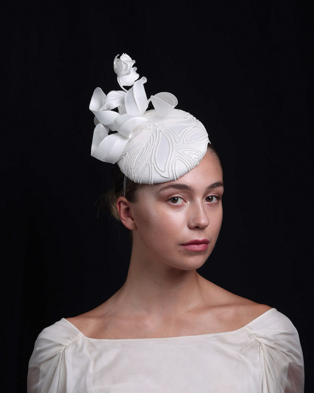 model is showcasing the side view of a button shaped fascinator. The base of the fascinator covered with sateen and contemporary elastic embroidery. Decorated with a sateen rose bud and swirled sateen leaves.
