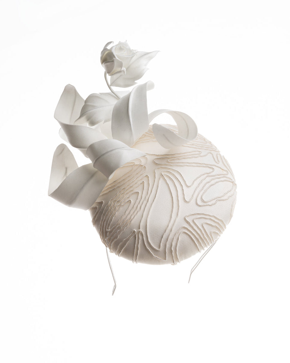a button shaped fascinator. The base of the fascinator covered with sateen and contemporary elastic embroidery. Decorated with a sateen rose bud and swirled sateen leaves.