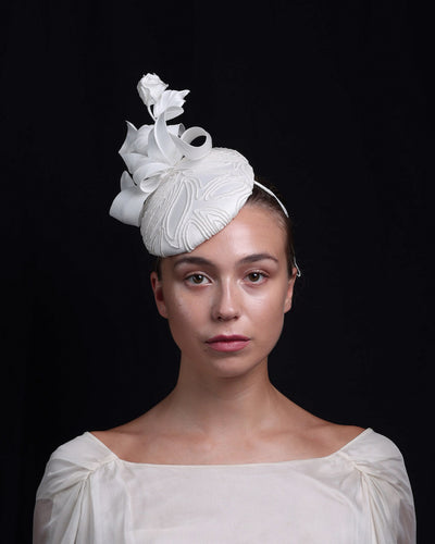 Model is showcasin the front view of a button shaped fascinator. The base of the fascinator covered with sateen and contemporary elastic embroidery. Decorated with a sateen rose bud and swirled sateen leaves.